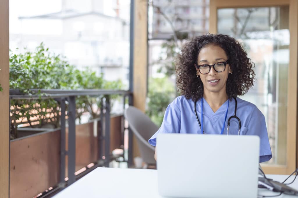 Nurse meeting with patient virtually using video call health technology Black female doctor uses a laptop computer to have a video call medical appointment with a patient. The medical professional is working from the patio of her home. Social distancing, Covid-19, telemedicine concepts.